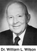 The image is a black and white photograph of an older man with gray hair, wearing a suit and tie, smiling slightly, and looking directly at the camera.